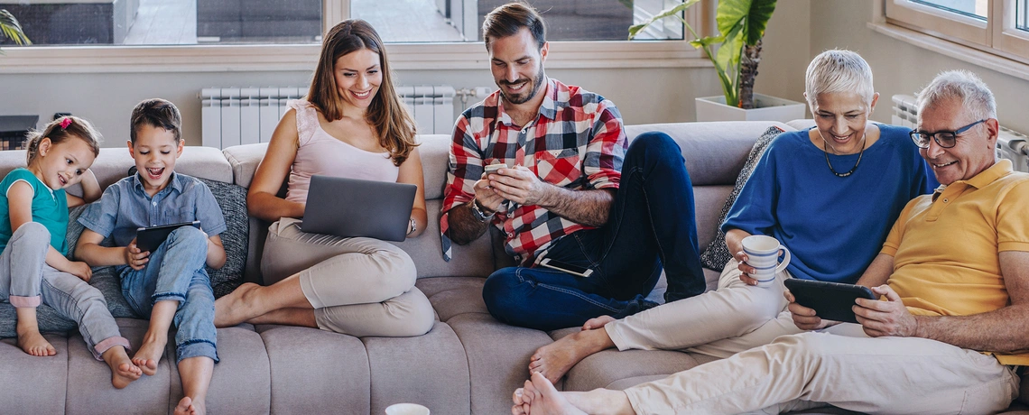 Drei Generationen sitzen gemeinsam auf dem Sofa und nutzen verschiedene digitale Geräte wie Tablet, Laptop und Smartphone.  